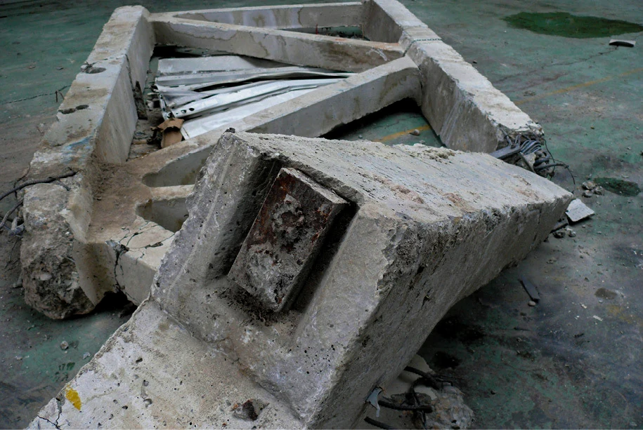 End of a collapsed segmented concrete truss lying on a hall floor, its broken head turned upward to show a rusted steel anchor plate embedded in the concrete