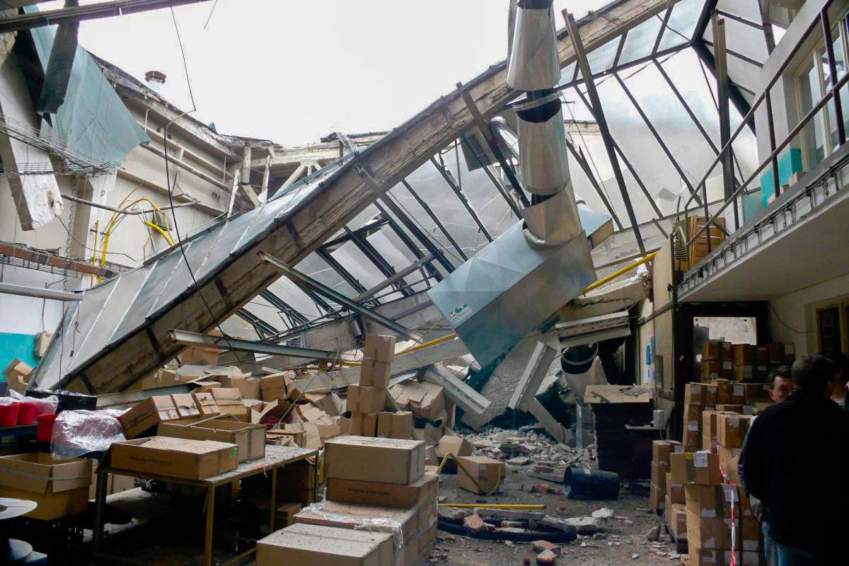 Interior of a warehouse after a roof truss collapse: a broken concrete lattice truss and torn roof panels hanging down over shelving and boxes, daylight coming through the gap