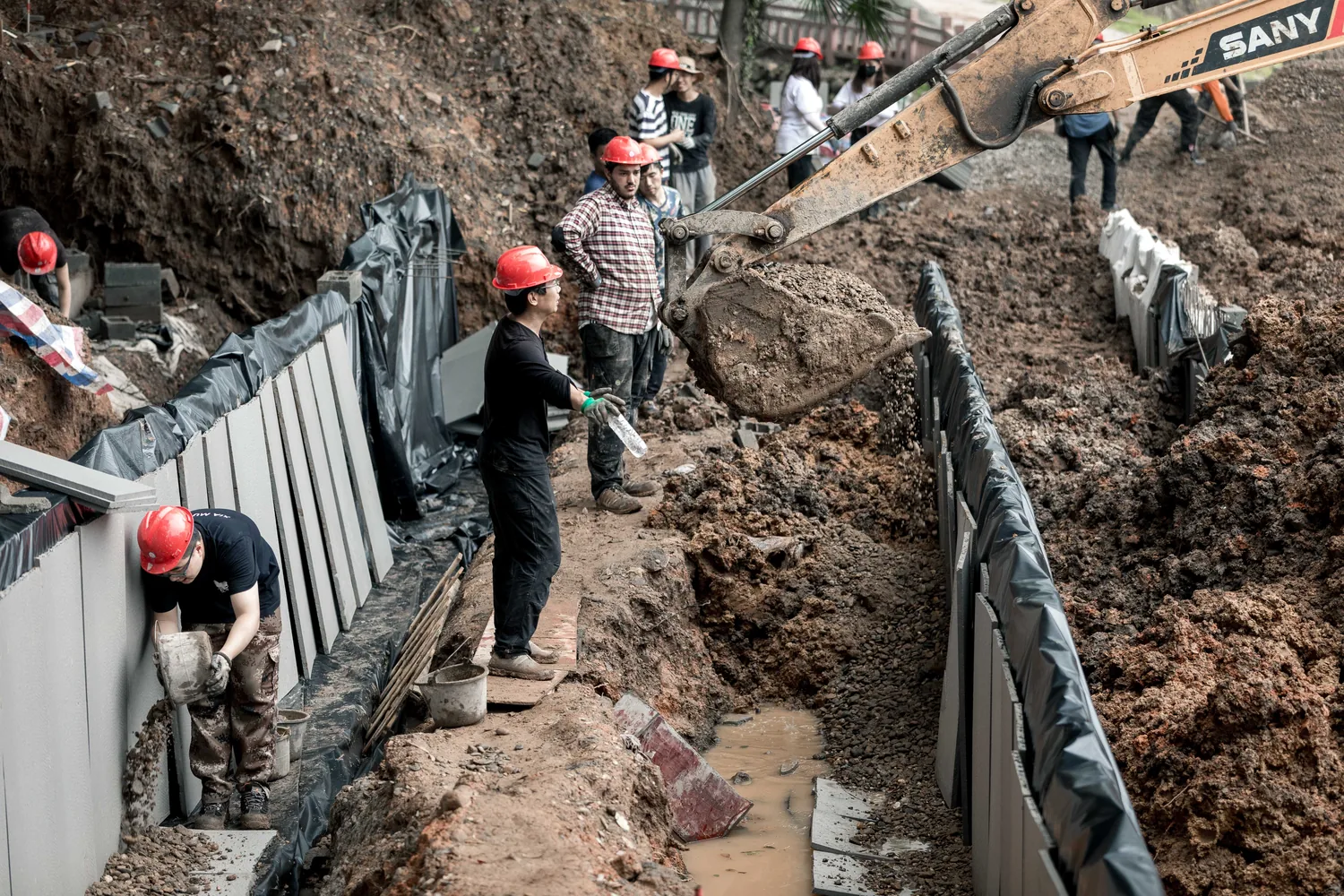 Students setting retaining wall blocks beside an excavator in a trench