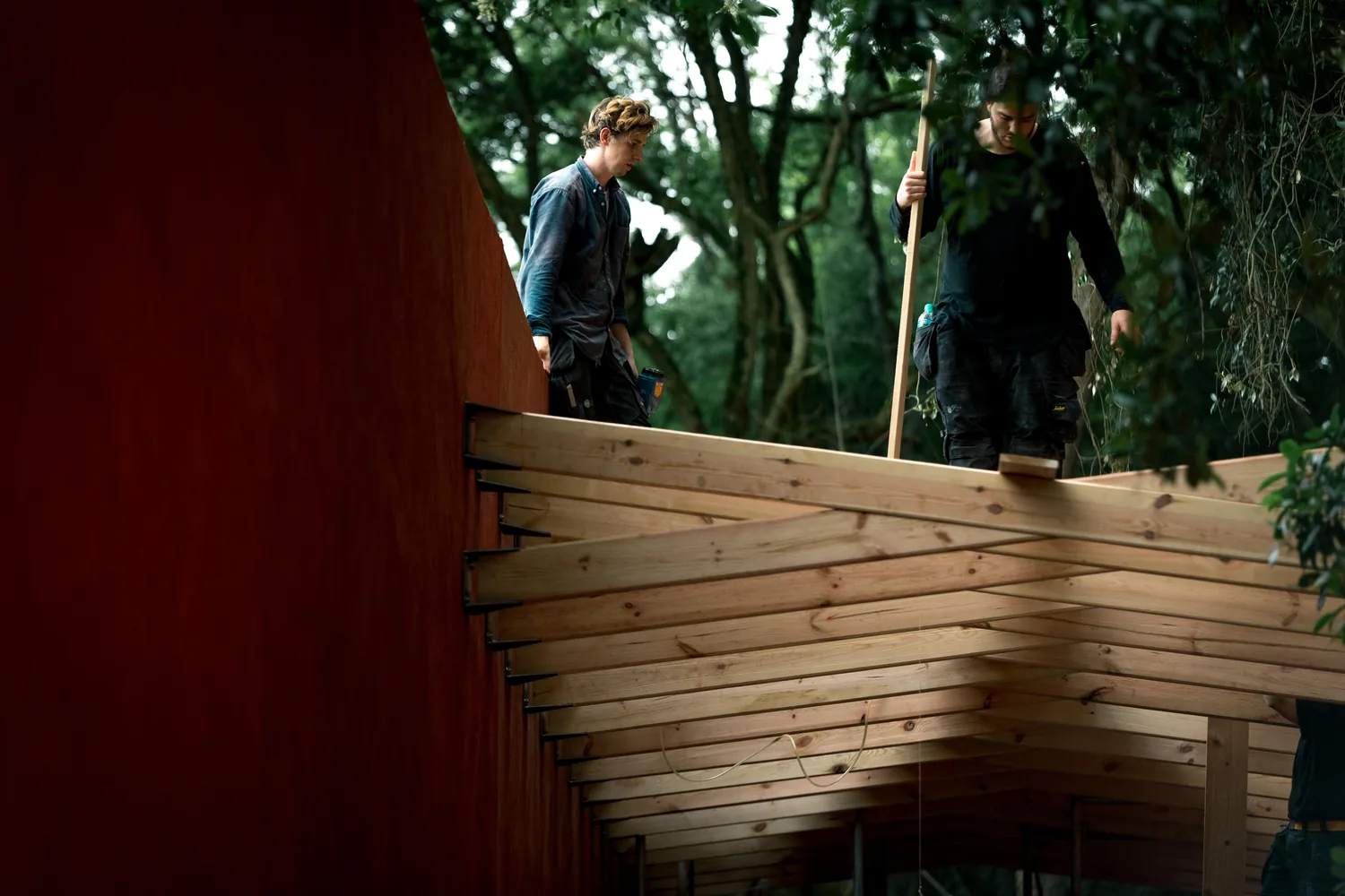 Two students assembling timber roof beams above a red wall